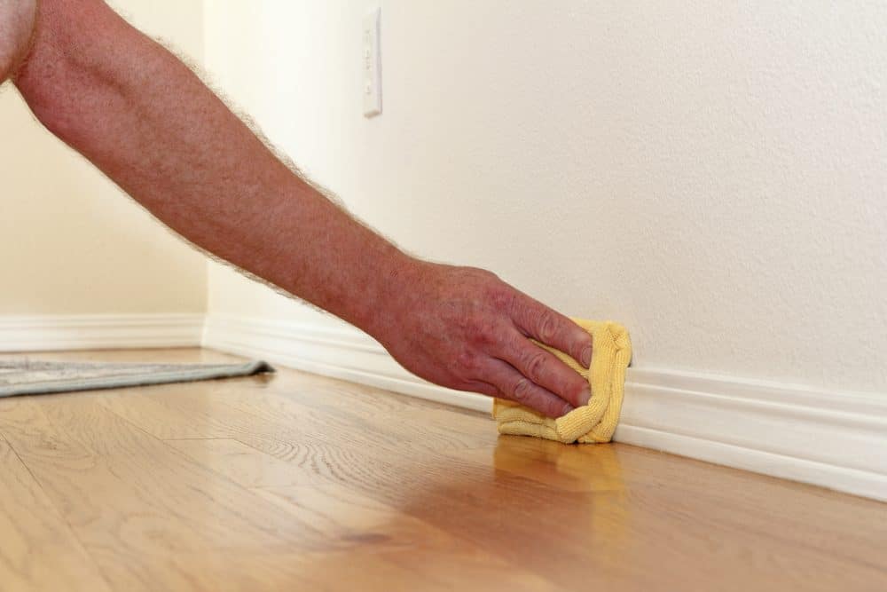 Male caucasian hand and arm seen wiping a folded yellow rag along the lower white wall trim near the wood floor to remove dust for a mold test.