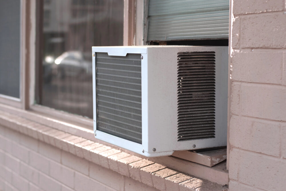 A window AC unit installed in a window surrounded by block wall construction painted pink. The window AC has mold.